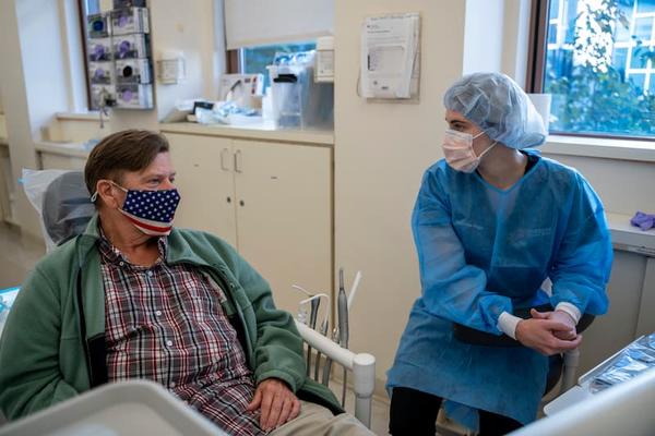 A veteran wearing an American flag facemasks sits in a chair speaking with a dentist.
