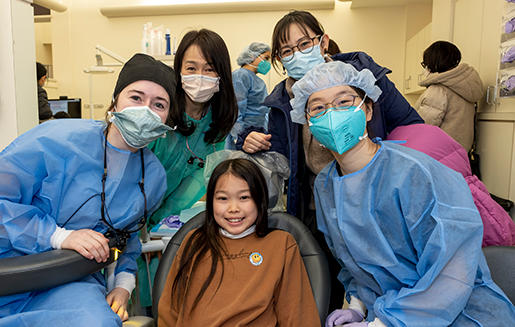 A group of dental professionals in scrubs and surgical masks poses with a smiling young girl in a dental clinic.