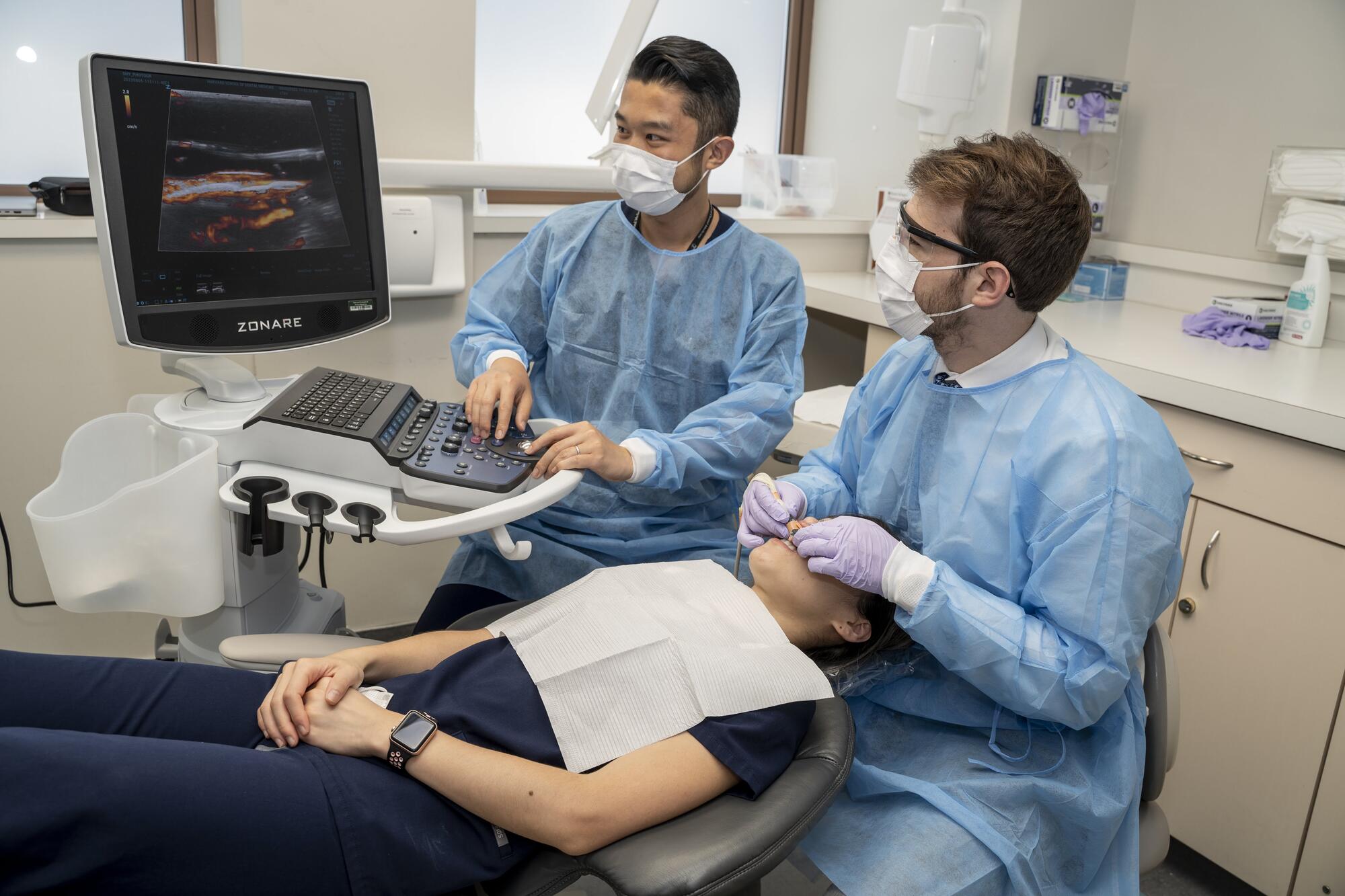Two dentists wearing blue scrubs working on patients mouth and looking at an ultrasound