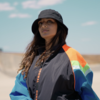 Person with long brown hair wearing a black bucket hat and multicolored windbreaker standing outside